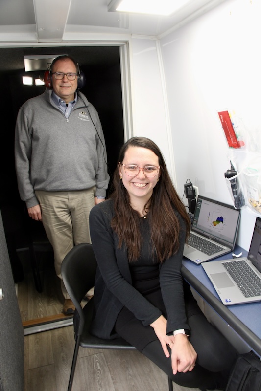 Alex Hustedt-Mai sits while Michael Heinz stands wearing headphones inside the APARC van.