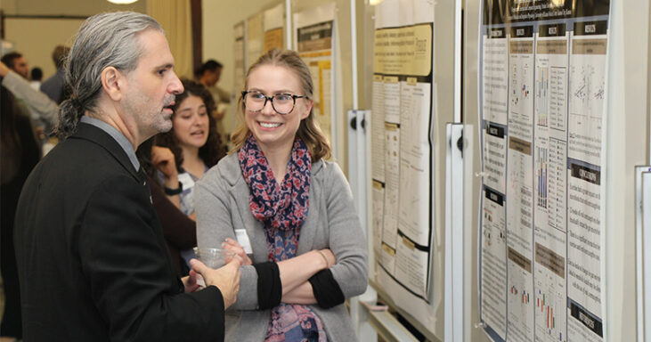 Two graduate students engage in friendly discussion in front of a Purdue-branded research poster.