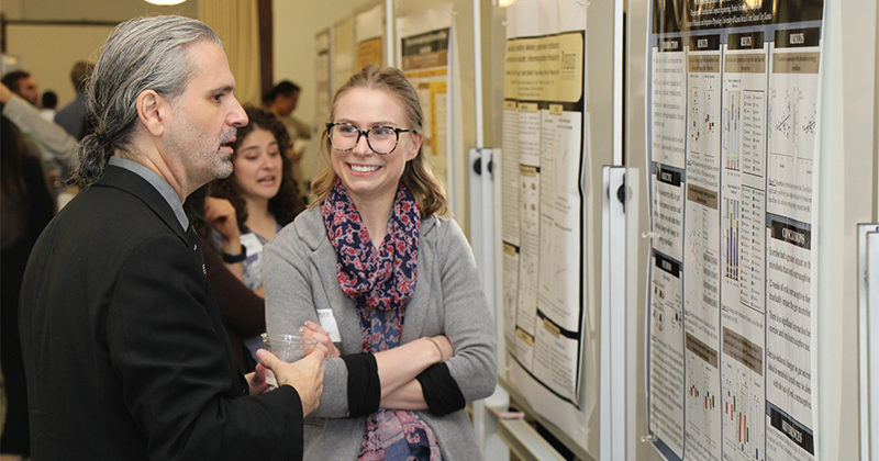Two graduate students engage in friendly discussion in front of a Purdue-branded research poster.