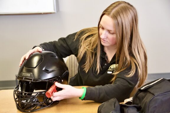 A graduate student starts to remove the face mask from a Purdue football helmet.