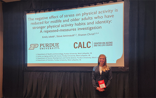 A young woman stands in front of a presentation screen, smiling. The presentation screen says, "The negative effect of stress on physical activity is reduced for midlife and older adults who have stronger physical activity habits and identity: A repeated-measures investigation."