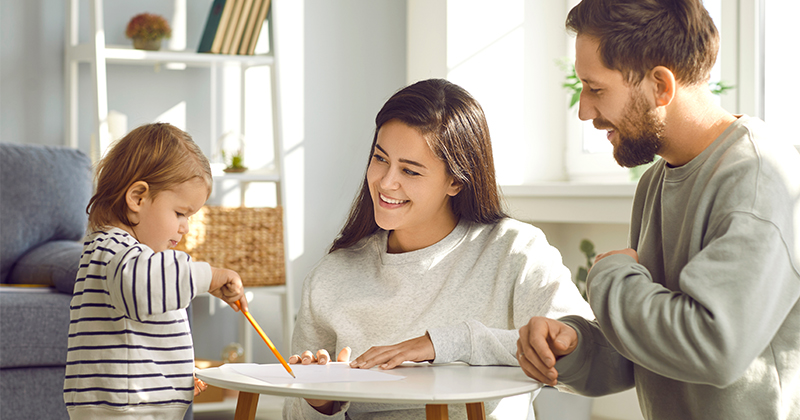 A young boy draws on a piece of paper with a pencil while his mother and father watch him, smiling.