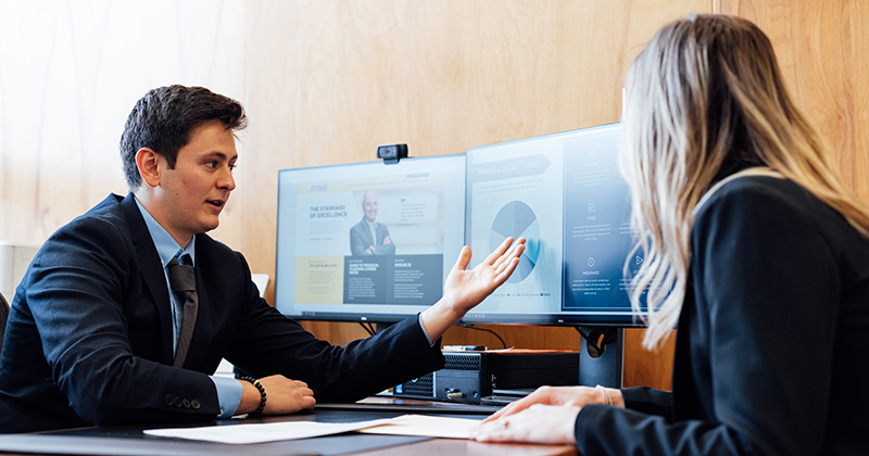 A student practices giving personal finance advice at a desk, pointing at computer screens.