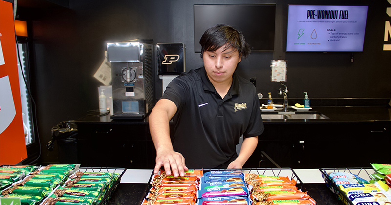Gabriel Pickart stands behind a counter containing a variety of nutritional bars, reaching to stock them.