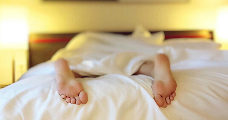 Feet hang off a bed, out of the blankets, with a white comforter covering the person laying down. 
