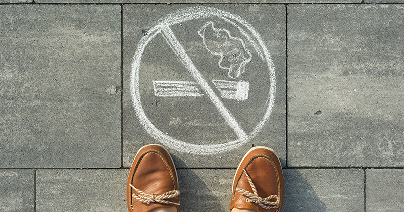 Feet stand on a sidewalk in front of a chalk drawing of a cigarette with a "no" symbol over it.