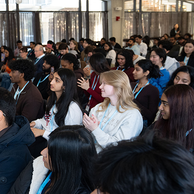 A crowd of people sit and clap for a speaker.