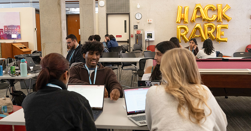 Four students sit at a table with laptops in front of them. Gold balloons spelling out "HackRare" appear in the background.