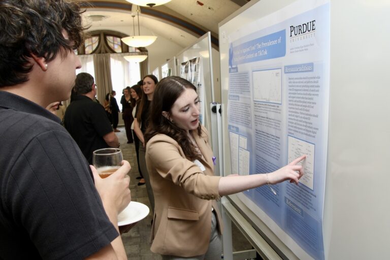Veronica Chavez talks to an onlooker in front of her research poster. She is pointing at a particular section on the poster.