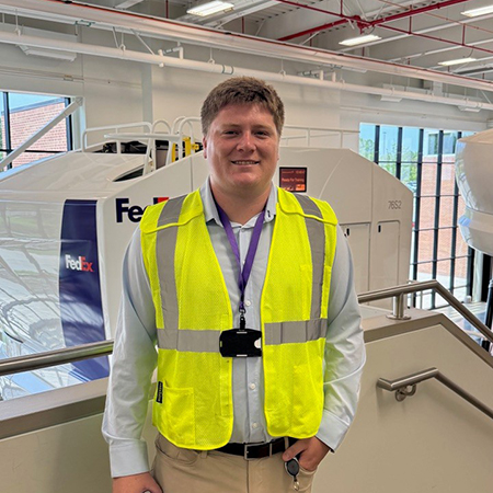 Odom stands in front of a FedEx flight simulator wearing a yellow safety vest during a tour of the FedEx World Hub.
