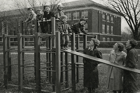 Young children stand on playground equipment with three women looking up at them.