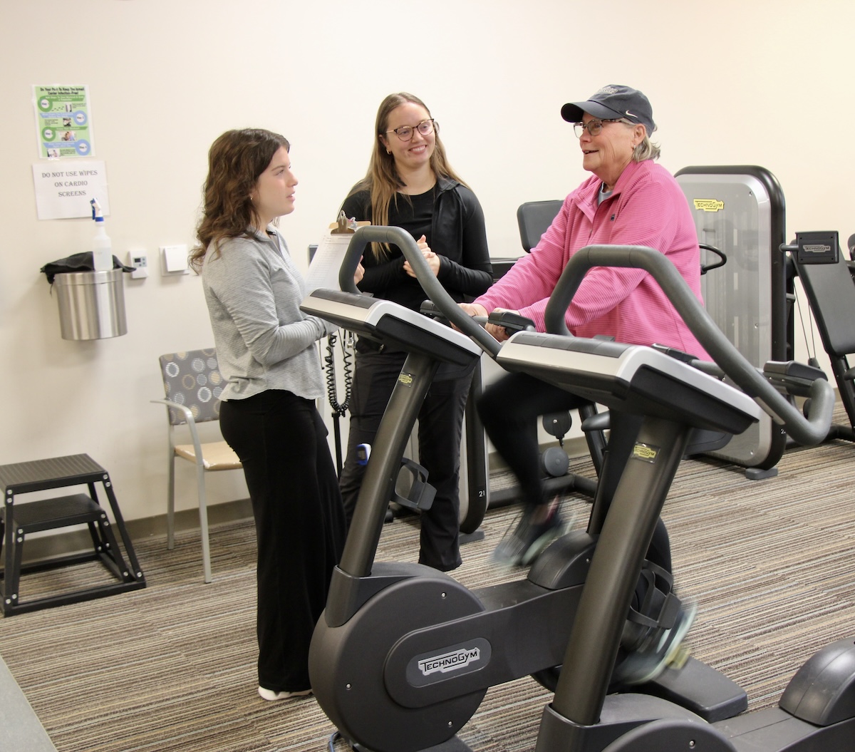 A woman pedals on an exercise bike while students observe.