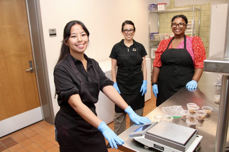A student researcher stands in the foreground while Annabel Biruete and a lab staff member stand in the background.