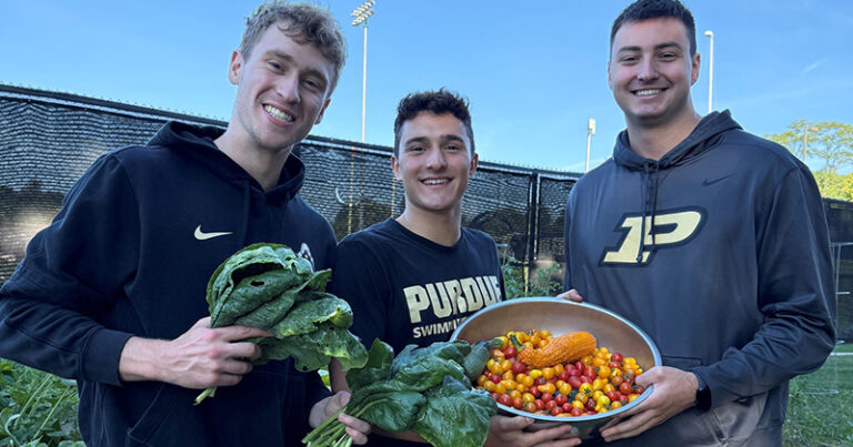 Students wearing Purdue gear hold up collected vegetables, smiling.