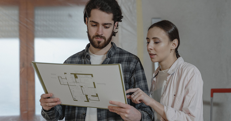 A couple looks at a house plan with one appearing to be correcting the other; both look unhappy.