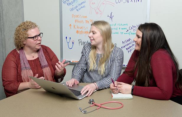 Two students work at a table with a professor. A whiteboard with writing about the human heart is behind them.