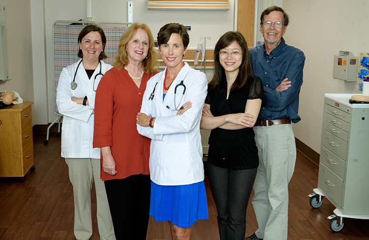 Four women and one man stand in a room that looks like a standard patient room in a hospital