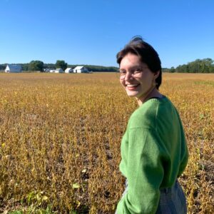 Molly Pfunder wearing glasses and a green sweater near a field of soy beans ready to be harvested.