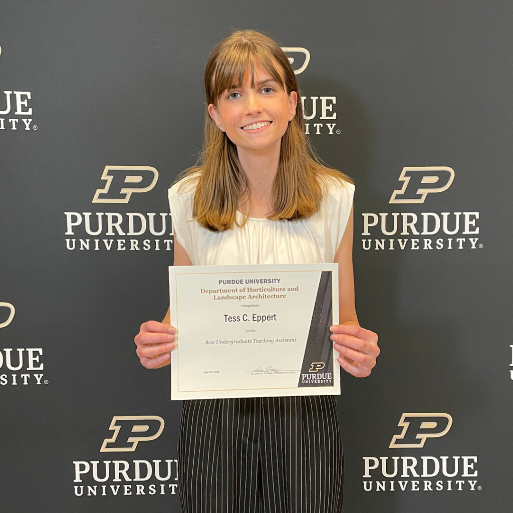 Tess Eppert stands in front of the Purdue backdrop holding her certificate.