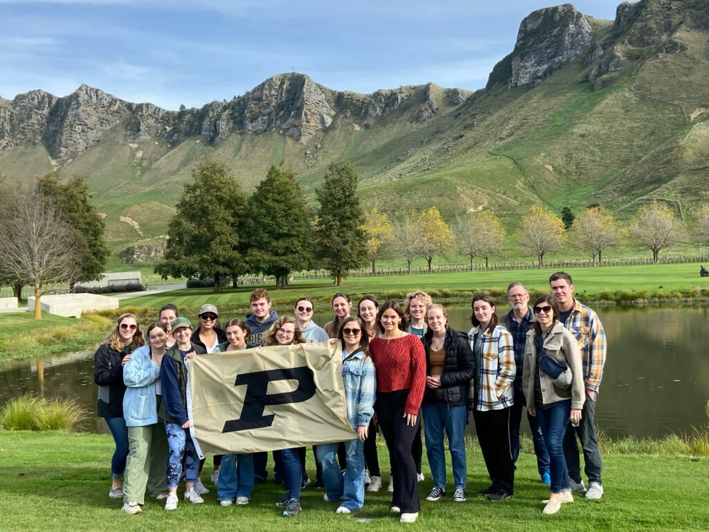 New Zealand Study Abroad group holding a Purdue flag outside with a pond and mountains in the background.