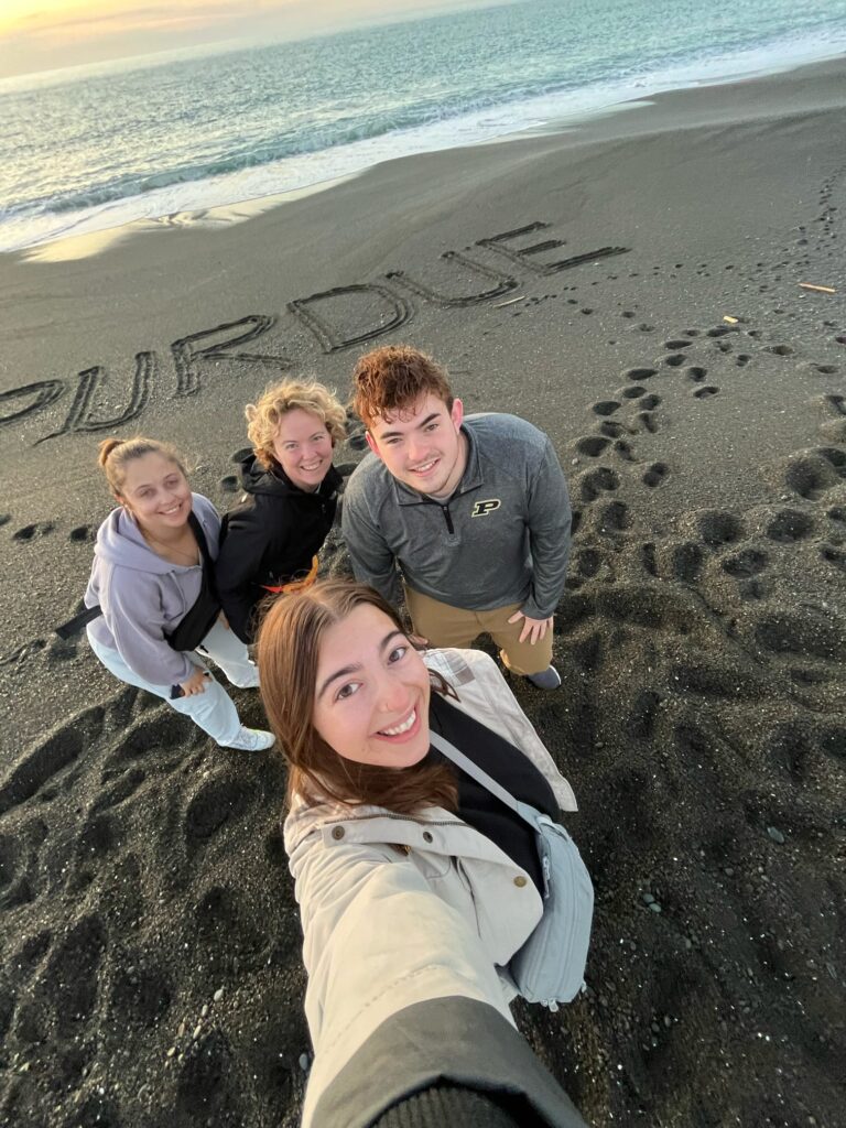 Four New Zealand Study Abroad students posing for a selfie at the beach with Purdue written in the sand.