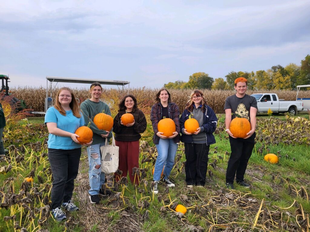 Six students standing in a field, all holding pumpkins.
