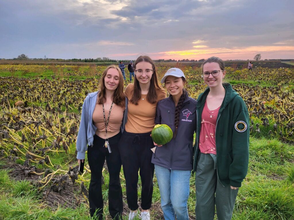 Four female students standing in a field, one holding a small green pumpkin.