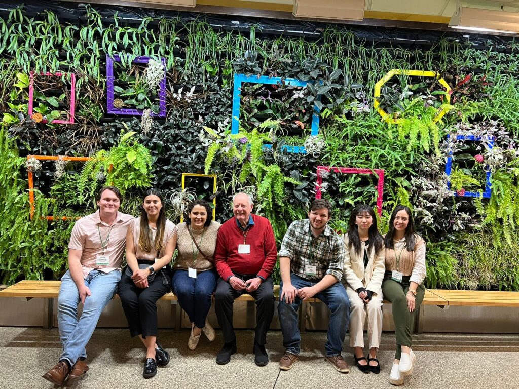Purdue-affiliated group of 7 people sitting in front of a plant wall.