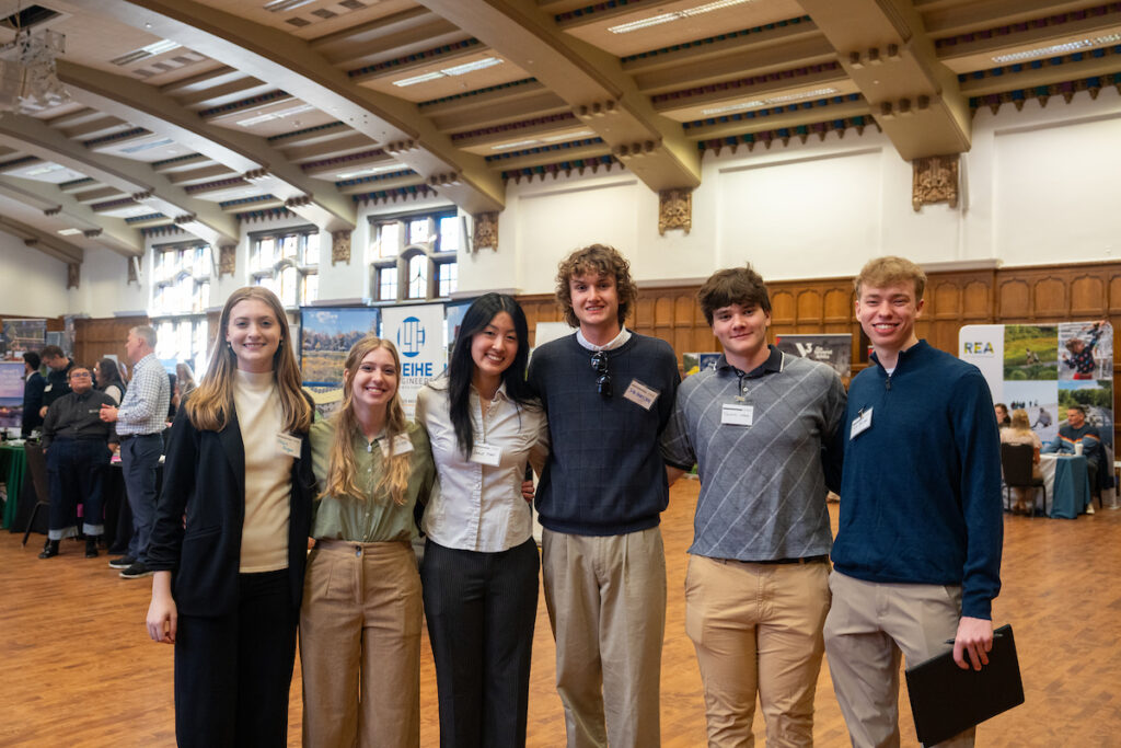 A group of landscape architecture students stand together in a large, wood-paneled hall during a career fair, with employer booths and informational displays visible behind them.