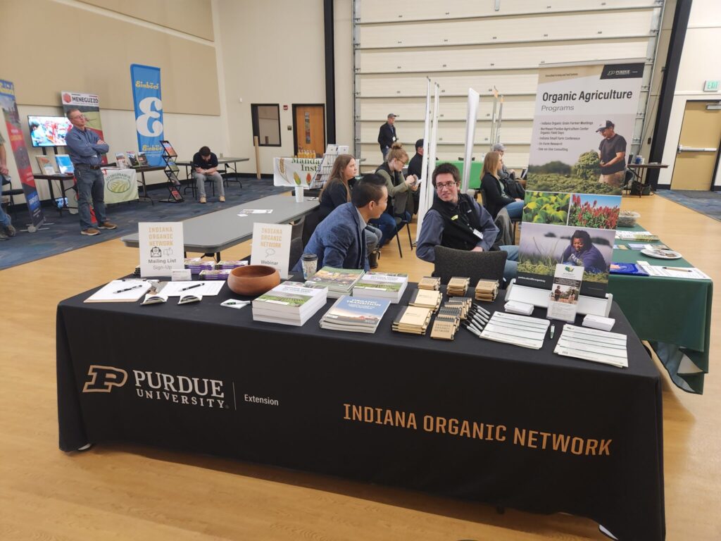 An Indiana Organic Network and Purdue University Extension exhibit table displays educational materials and publications, with attendees seated nearby during the meeting.