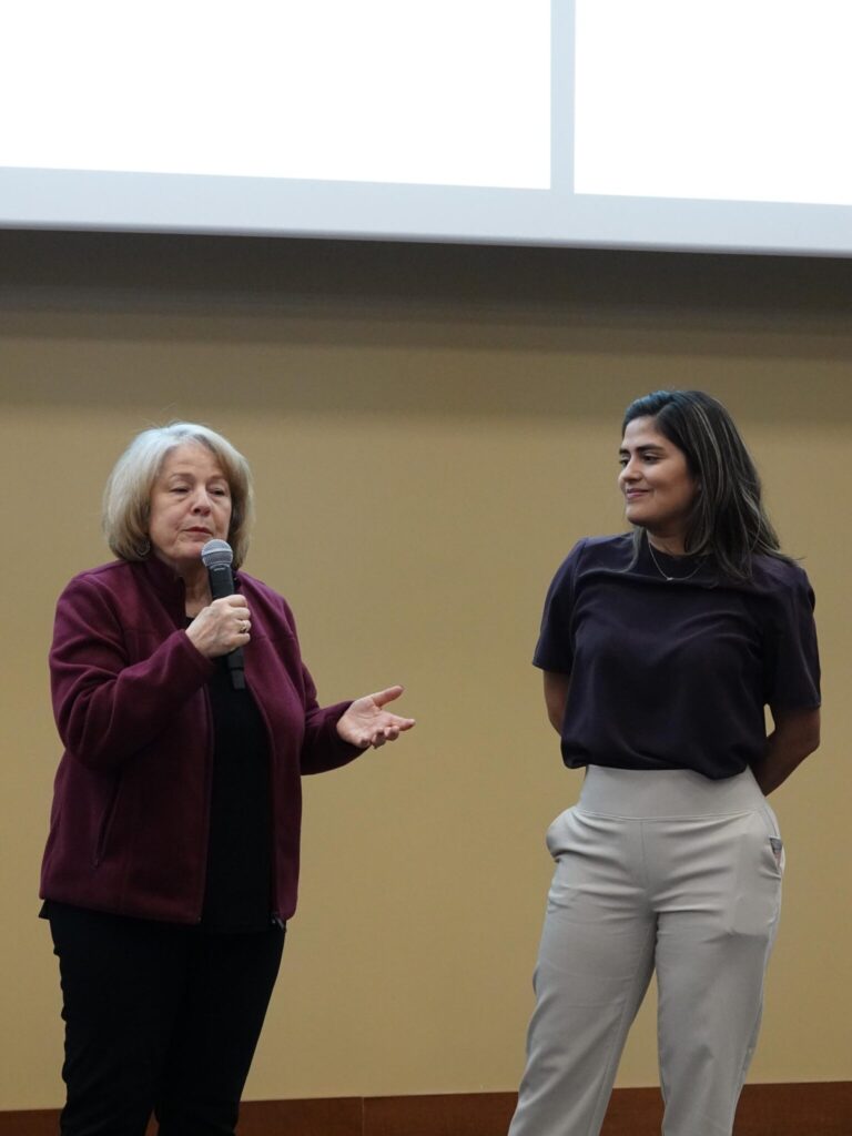 One person speaks into a microphone during an award ceremony while the award recipient stands nearby listening, both positioned at the front of a lecture hall beneath a projection screen.