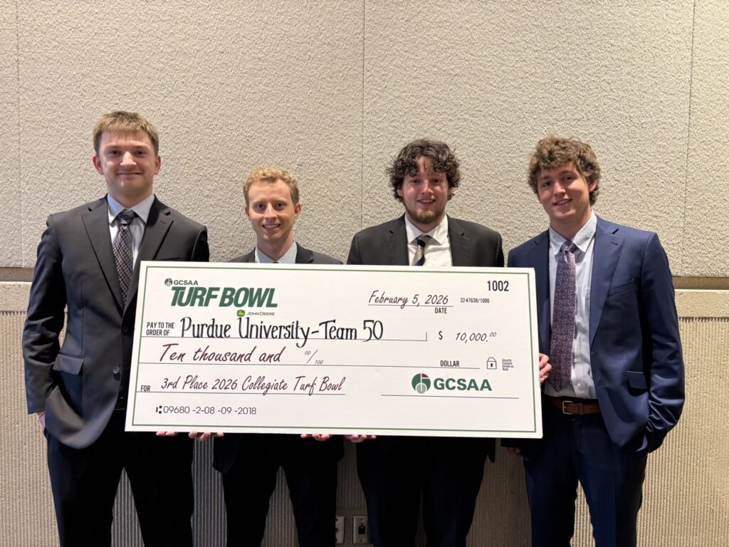 Four Turf Science students standing indoors holding a large ceremonial check for a Turf Bowl competition award