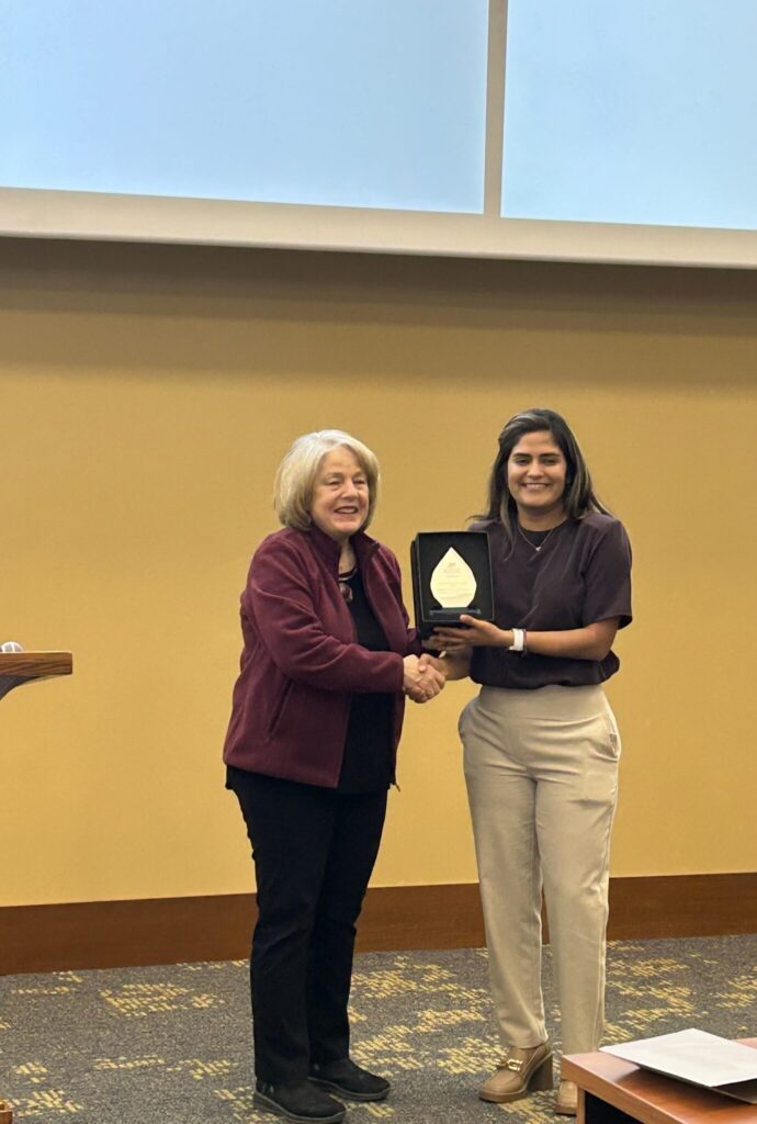 Two people stand in a lecture hall during an award presentation, shaking hands as one presents a plaque, with a projection screen visible overhead.