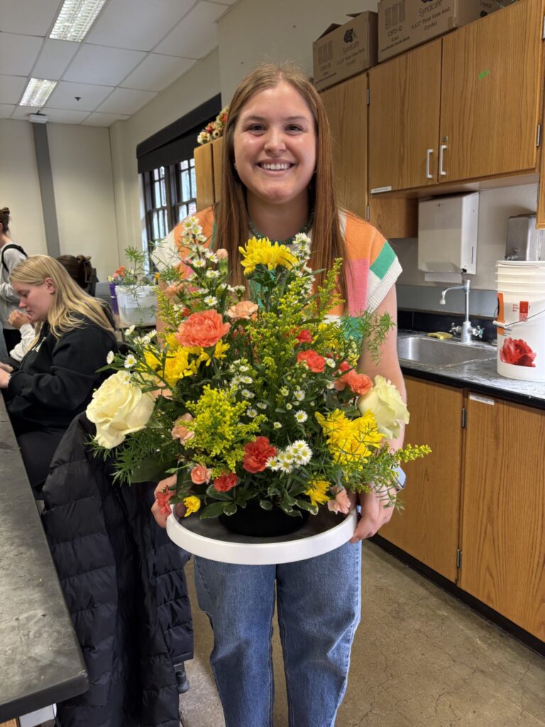 Student holding a completed round floral centerpiece with yellow, orange, white, and green flowers in a classroom setting.
