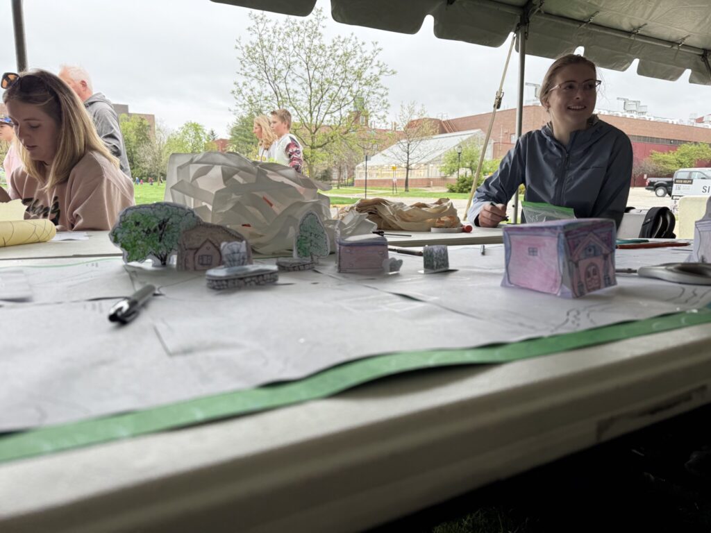 Participants sit under a tent working with paper models to build small landscape scenes, with drawn buildings and trees arranged on a tabletop.