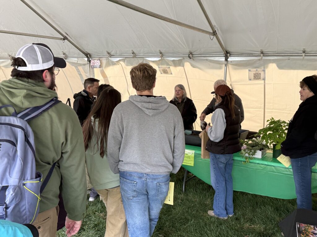 Master Gardener volunteers demonstrate container gardening to a group gathered under a tent, with plants and materials arranged on tables.