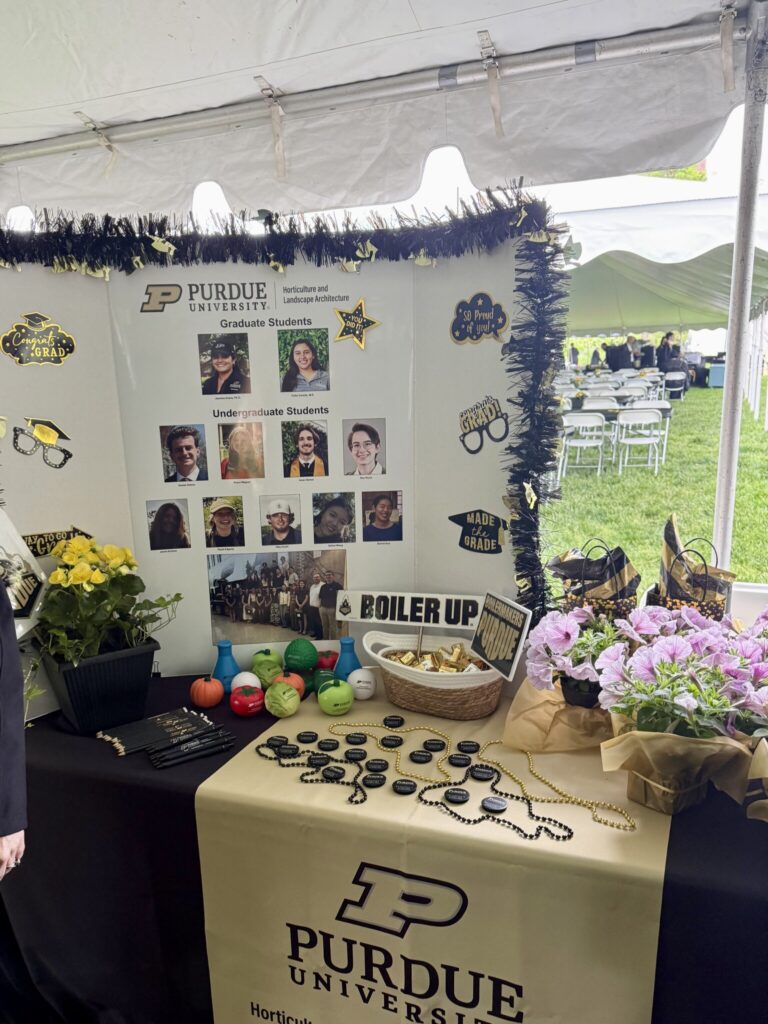Purdue HLA display table under a tent featuring graduate and undergraduate student photos, “Boiler Up” signage, small promotional items, and potted flowers.
