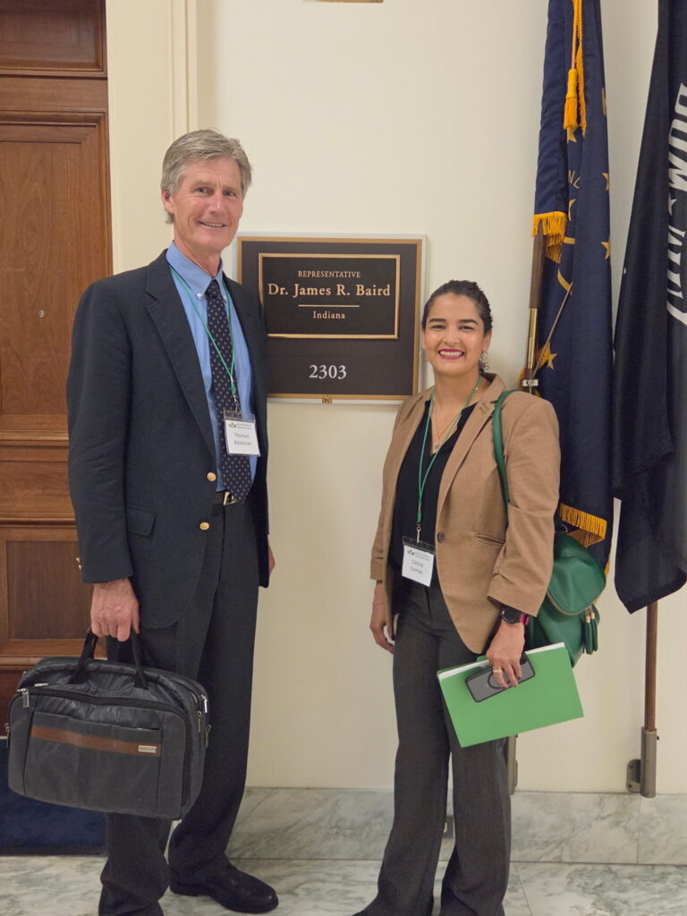 Two attendees stand in a hallway beside a plaque reading “Representative Dr. James R. Baird, Indiana,” holding folders and a briefcase.