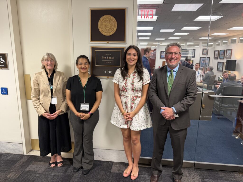 Four attendees stand in a hallway outside an office with a plaque reading “Senator Jim Banks, Indiana,” with a meeting visible through the glass door behind them.