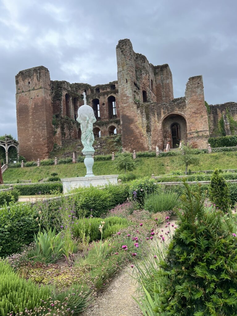 Ruins of Kenilworth Castle rise behind a formal garden with flowering plants and a central statue holding a globe.