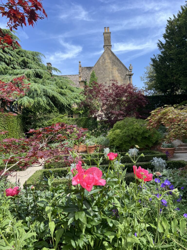 Hidcote garden courtyard with colorful flowers in the foreground, clipped hedges, and a stone house under a bright sky.
