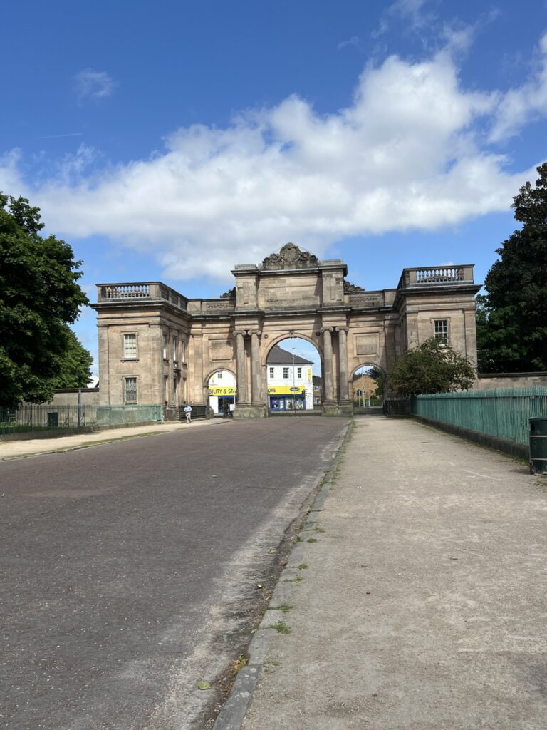 Grand stone park entrance arch at Birkenhead Park with a wide roadway passing beneath and trees on either side.