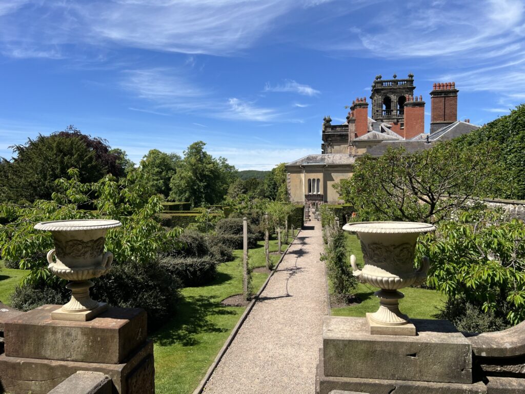 Formal garden at Biddulph Grange with a straight gravel path, large urn planters, trimmed hedges, and a historic building beyond.