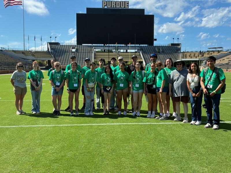 Group of 4-H'ers and staff wearing matching green T-shirts standing on the field inside Purdue’s football stadium, with a large scoreboard and “PURDUE” visible in the background under a blue sky.