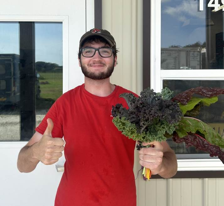 Raffi Lindsay standing outside a building holds a bouquet of leafy greens, including kale and Swiss chard, while giving a thumbs-up gesture.