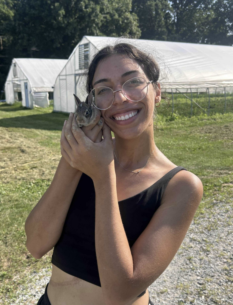 Kayla Quintero holding a young rabbit beside their face while standing in front of greenhouse structures on a farm.