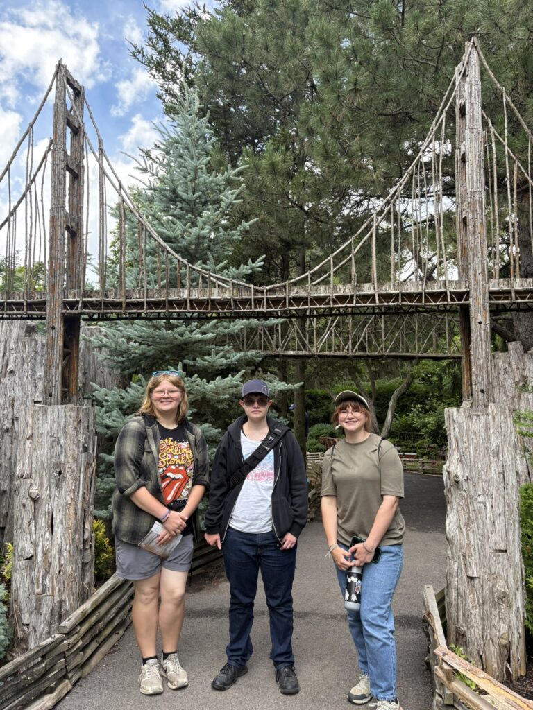 Three HLA student gardeners stand beneath a rustic wooden suspension bridge display in the Chicago Botanic Garden’s model railroad garden.