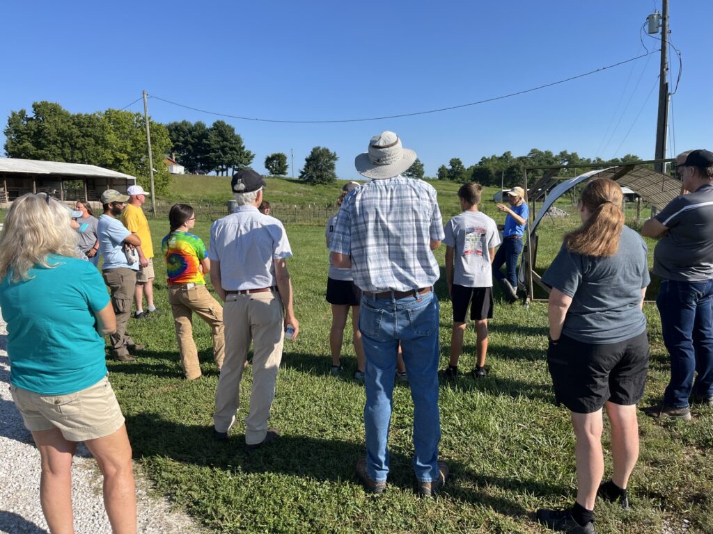 A group of participants gathers outdoors in a pasture while a presenter leads a discussion near a portable livestock shelter.