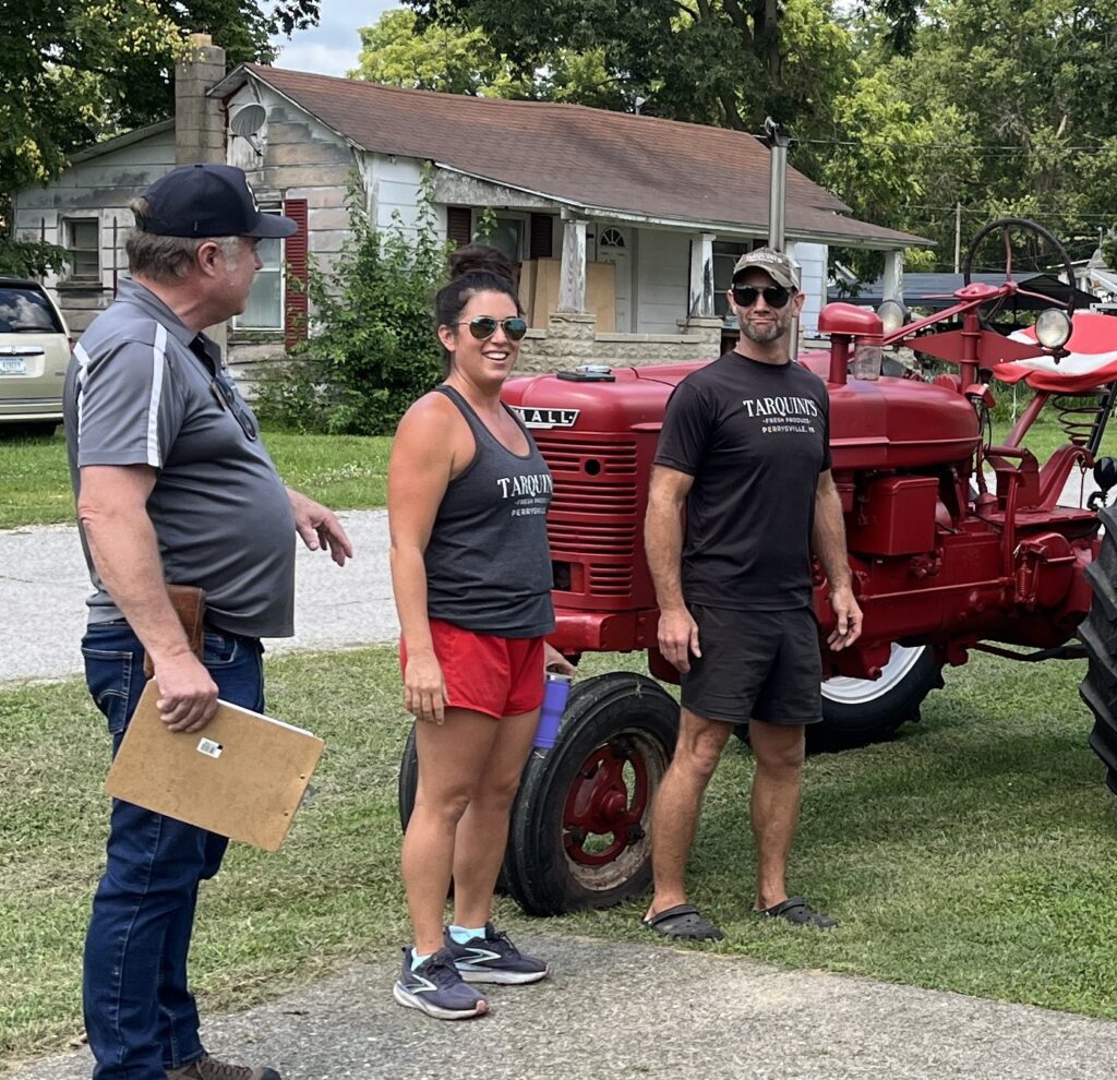 Three people stand beside a restored red vintage tractor in a grassy residential area, listening and talking during an outdoor farm-related event.