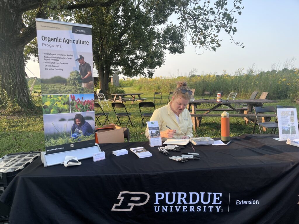 Purdue Extension Organic Agriculture information table at an outdoor event, with brochures, pens, and a banner promoting organic agriculture programs beneath large shade trees.